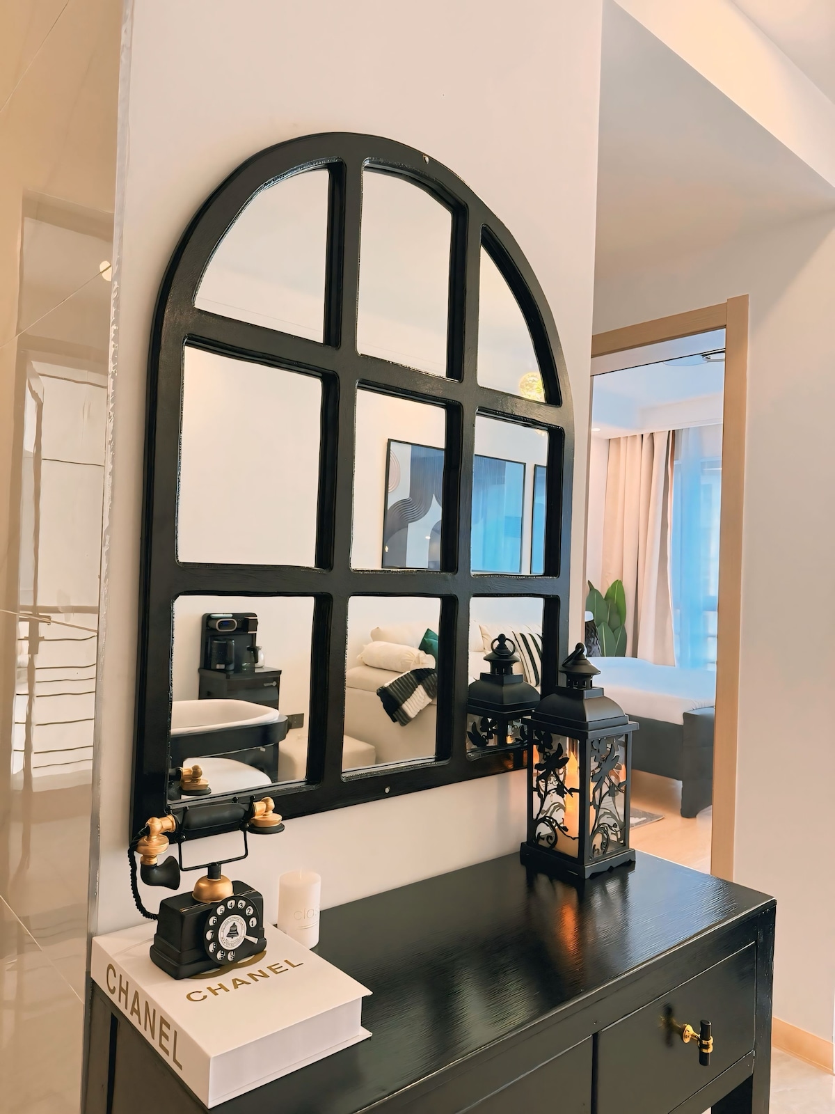 A stylish entryway features a large arched mirror framed in black, reflecting light and space. A vintage-style telephone rests atop a small side table, accompanied by a decorative lantern. This setup enhances a welcoming atmosphere within the apartment.