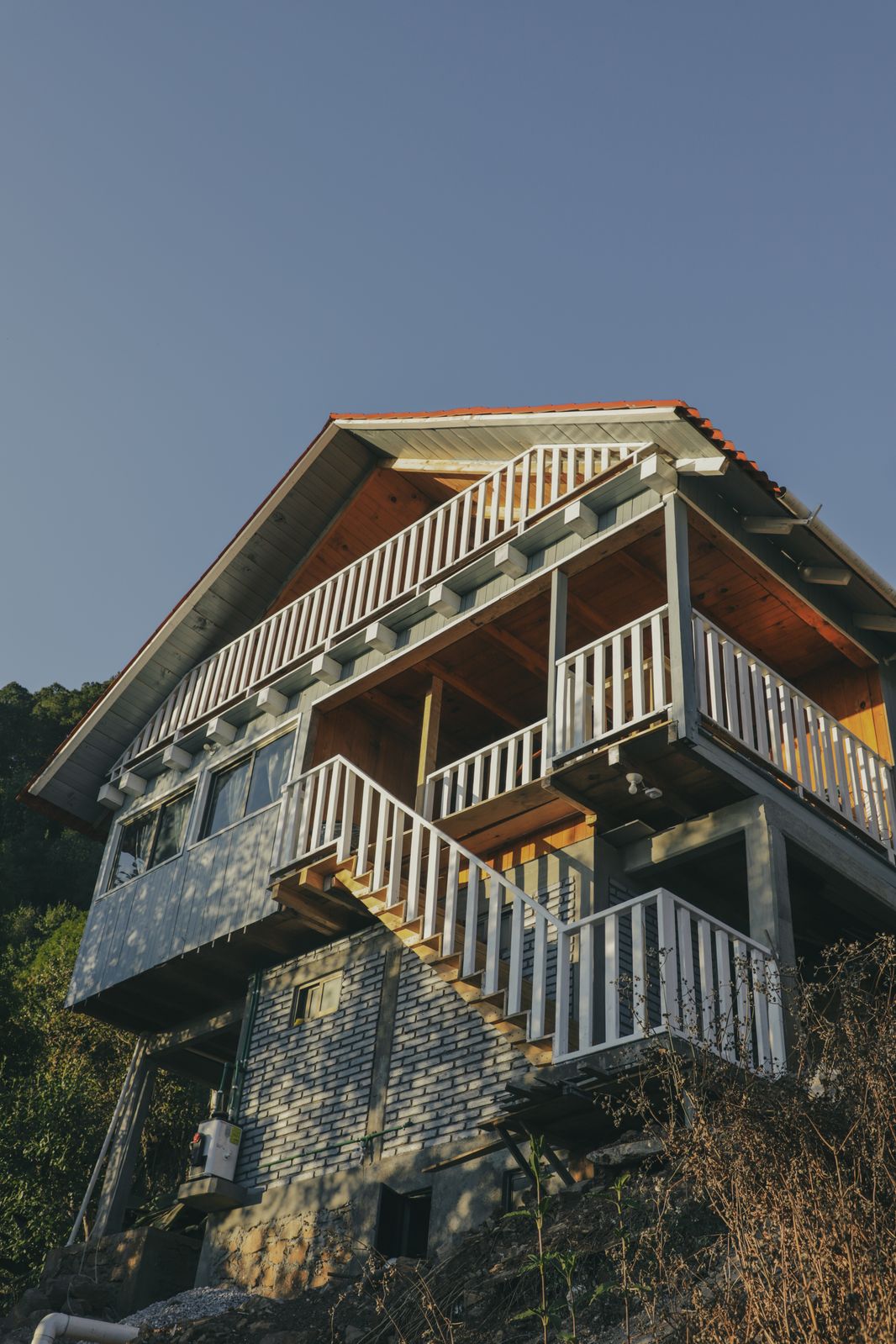 A two-story cabin is observed from below, showcasing a wooden exterior with a red-tiled roof. Multiple balconies with white railings are visible. A stairway leads to the entrance, surrounded by natural greenery.