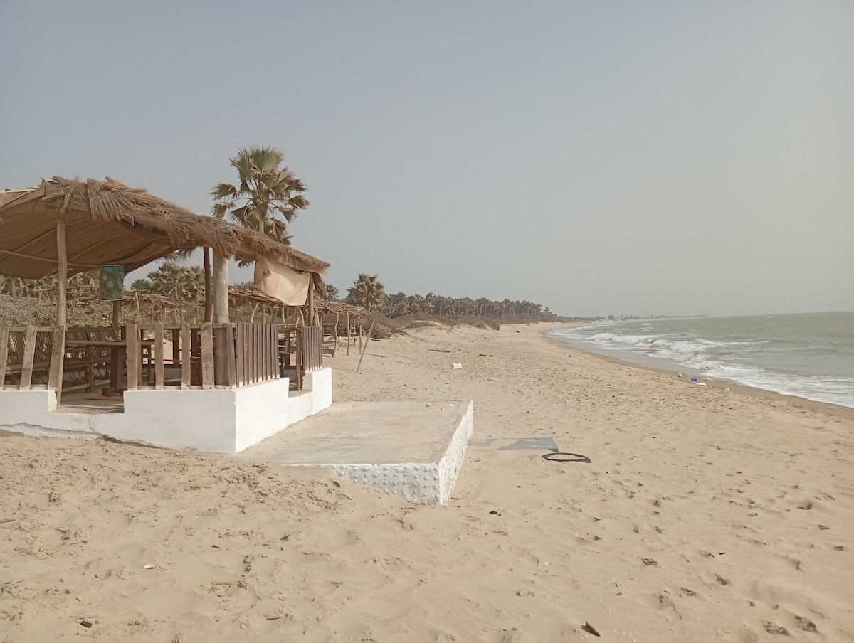 A sandy beach stretches along the shoreline, accompanied by gentle waves. A shaded seating area is positioned on a raised platform, featuring wooden furnishings. Palm trees are visible in the background, providing a natural setting. The scene conveys a sense of tranquility by the ocean.