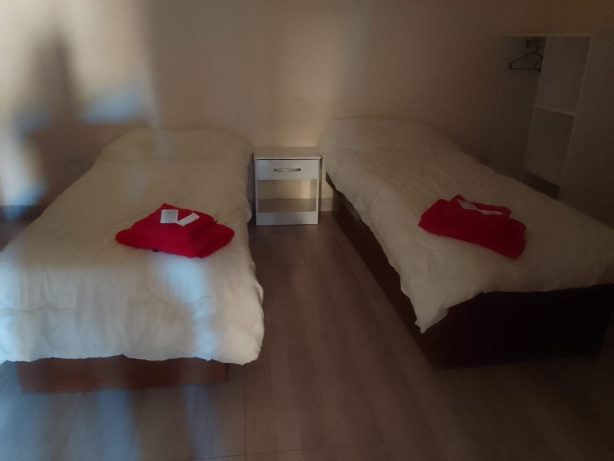 Two separate beds are positioned within a softly lit room, each adorned with white bedding and a neatly folded red blanket. A small nightstand sits between the beds, providing a practical space for personal items. A simple shelf is visible in the corner.