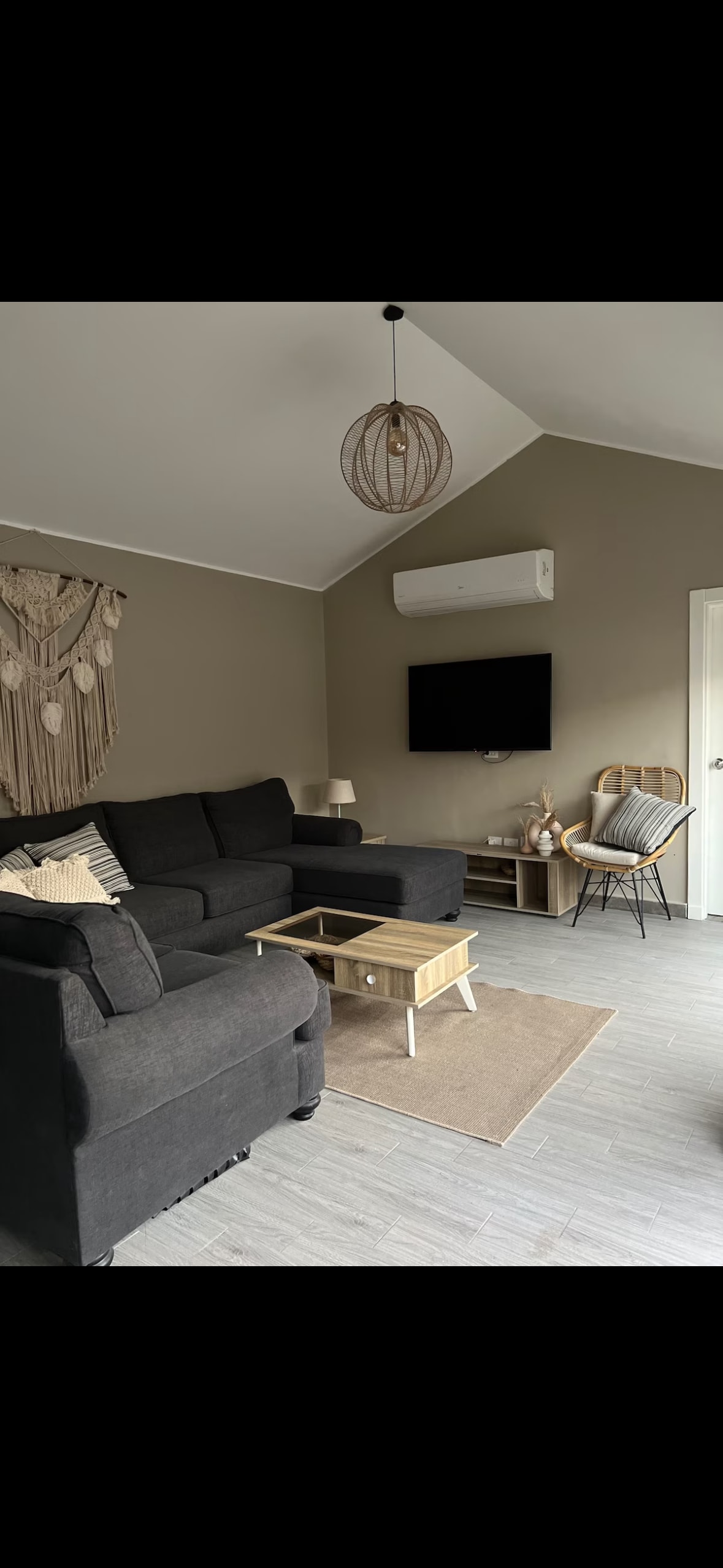 A comfortable living area is portrayed, featuring a dark sectional sofa accompanied by a light-colored coffee table. A flat-screen television is mounted on the wall, and a woven light fixture adds texture to the soft-toned room. A small rug anchors the seating area.