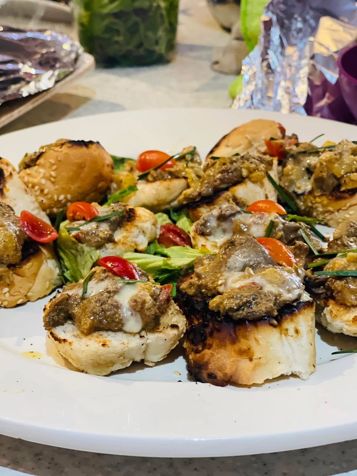 An assortment of toasted bread slices topped with a flavorful mixture, featuring herbs and pieces of meat. Fresh cherry tomatoes and leafy greens are arranged on the plate, adding a vibrant touch to the presentation.