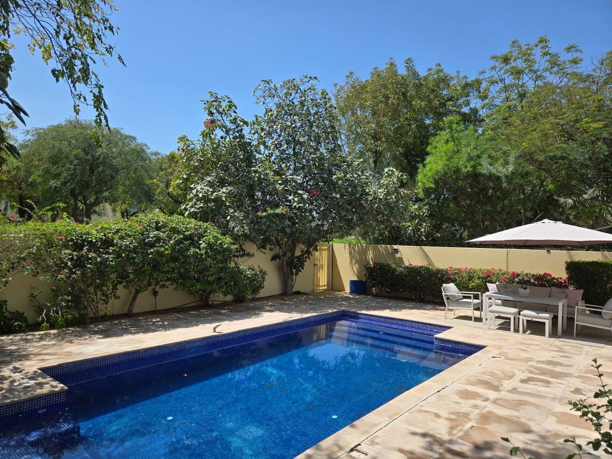 A private pool is surrounded by lush greenery and flowering plants, providing a serene outdoor retreat. Comfortable lounge chairs and a shaded dining area are situated beside the pool, offering space for relaxation and meals in a tranquil setting.