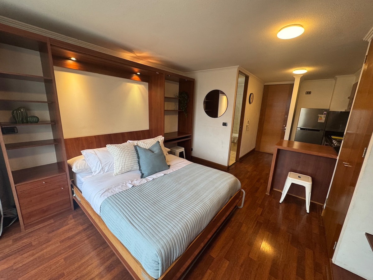 A studio apartment features a wooden bed frame with a soft blue bedspread. Shelving units are positioned beside the bed, and a small desk is located across from it. The space is illuminated by a ceiling light, contributing to a warm ambiance.