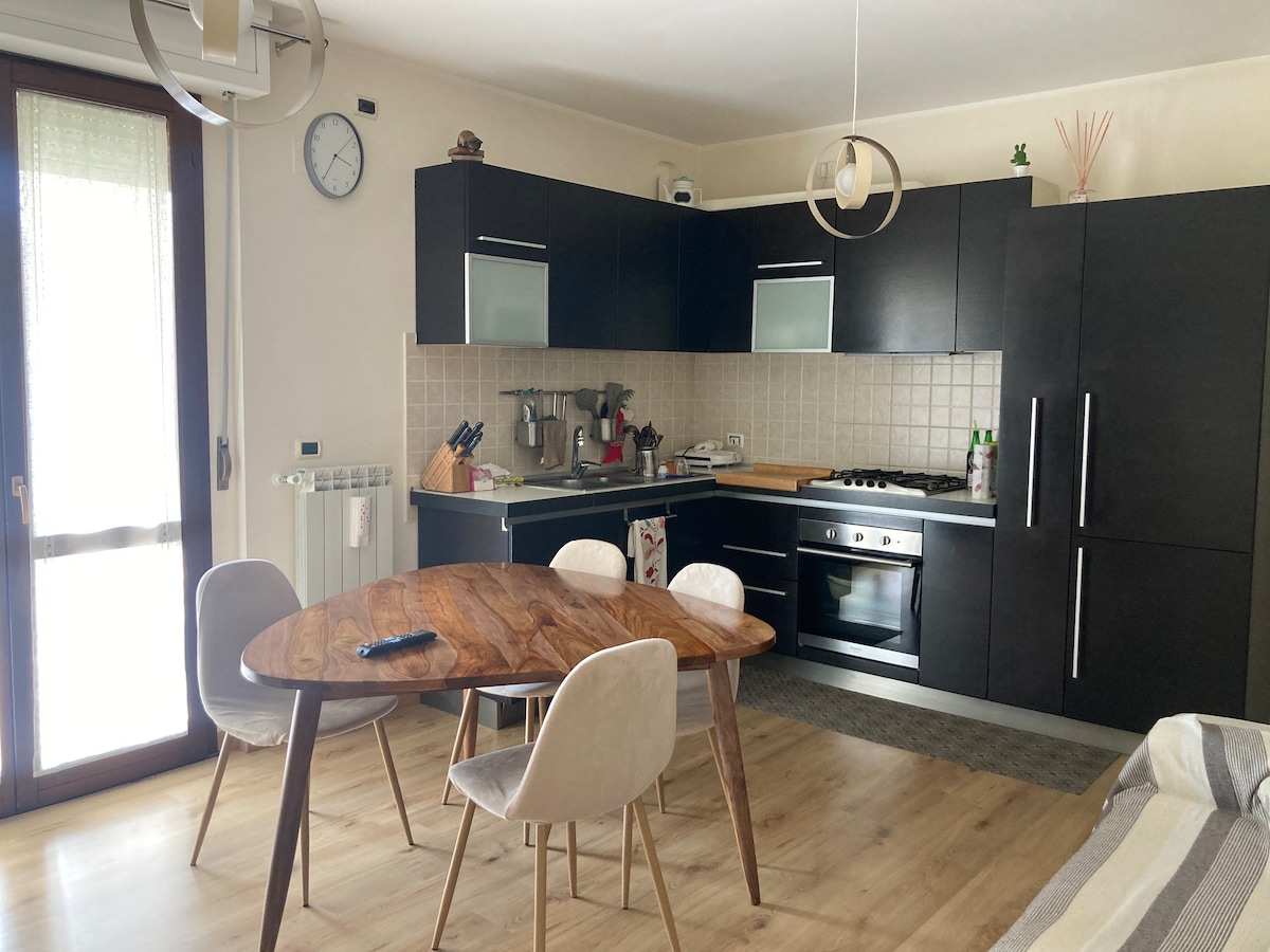 A modern kitchen with dark cabinetry and stainless steel appliances is complemented by a round wooden dining table surrounded by four light-colored chairs. Natural light enters through a glass door leading to a balcony, enhancing the inviting space.