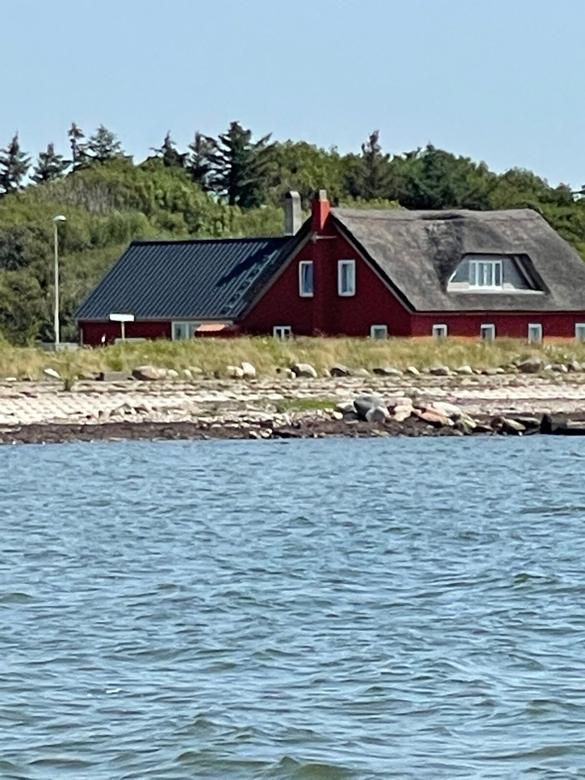 A red building with a thatched roof is visible near the shoreline, surrounded by greenery. The structure features a sloped roof and several windows, offering views of the adjacent water and landscape. The scene captures a sense of tranquility by the coast.