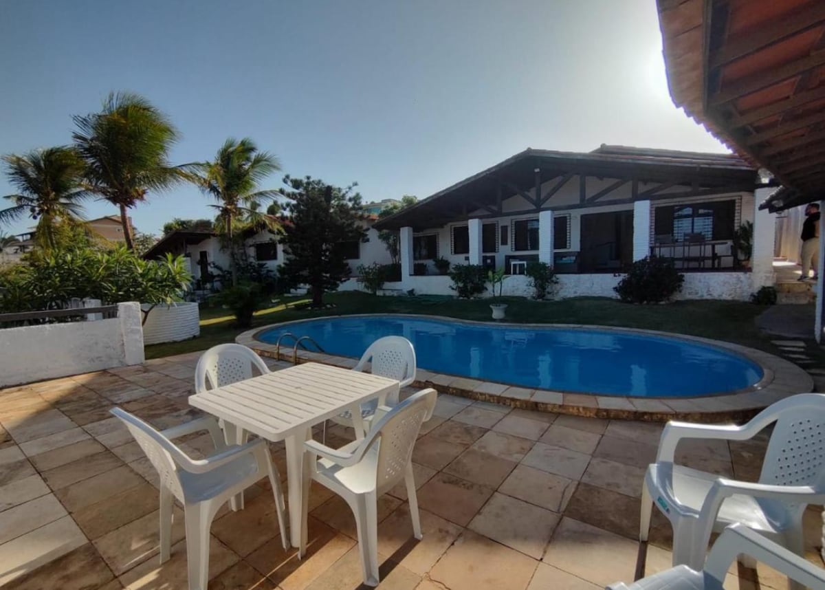 A spacious outdoor area features a rectangular pool surrounded by tropical greenery. Several white plastic chairs are arranged around a table on a tiled patio. The house, with a wide porch and multiple windows, is visible in the background under clear blue skies.