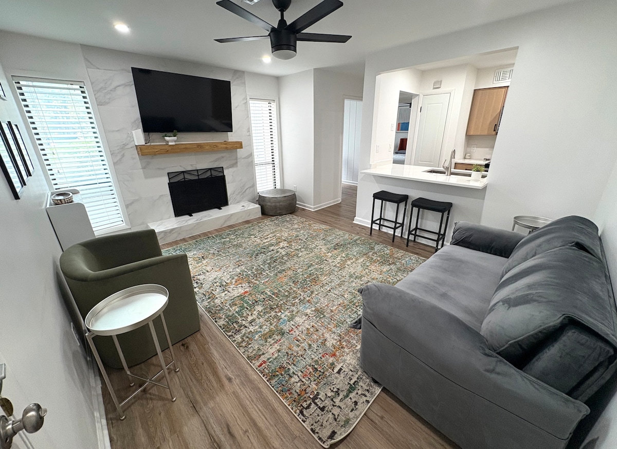 The living area is centered around a stylish gray sofa and two accent chairs. A large area rug adds color to the space, with windows allowing natural light. A wall-mounted TV is positioned above a wooden mantel, while a small round table is placed nearby.