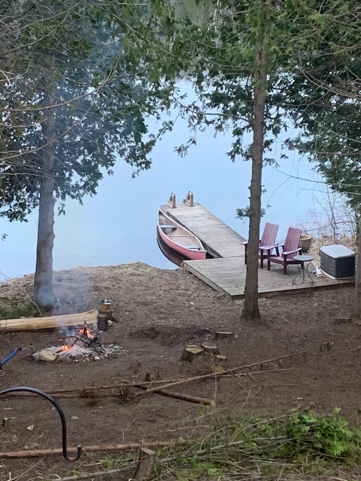 A serene lakeside scene captures a red canoe resting on the edge of the water, alongside a wooden dock. A small fire pit with burning logs is visible, surrounded by natural foliage. Two chairs are positioned nearby, creating a cozy space for relaxation.
