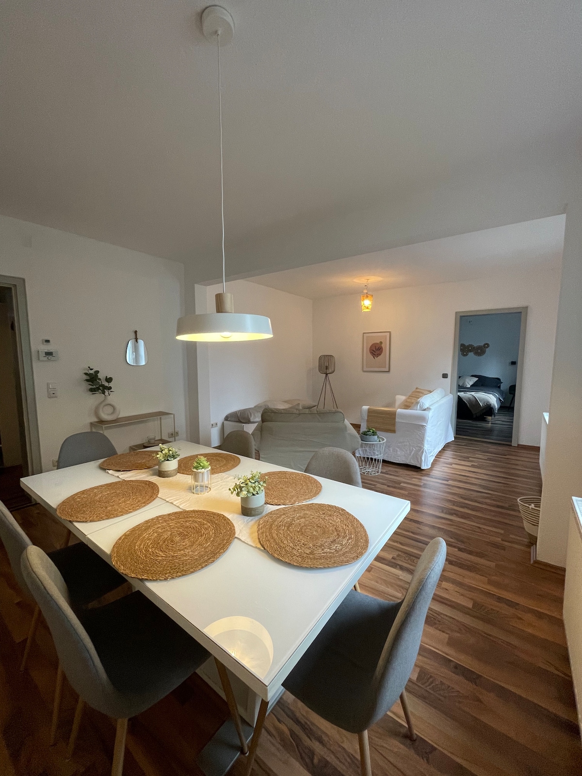 A well-lit dining area features a white table with five round placemats and six upholstered chairs. A pendant light hangs overhead, illuminating the space. In the background, a cozy living area is visible, showcasing a soft sofa and a glimpse of a bedroom.