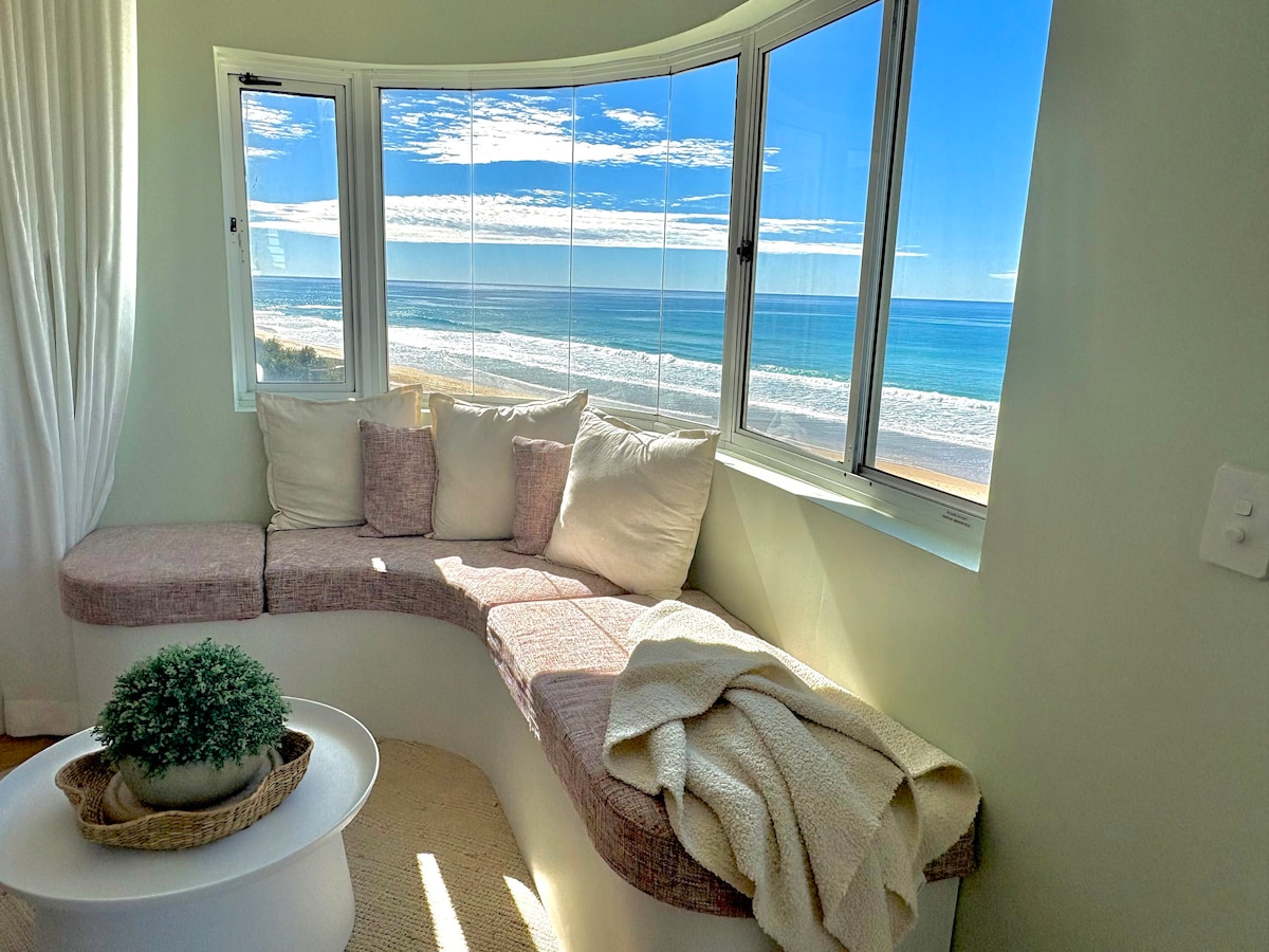 A curved seating area features plush cushions and a soft blanket draped casually. Large windows provide expansive views of the beach and ocean, allowing natural light to brighten the space. The serene coastal scenery creates a calming backdrop for relaxation.