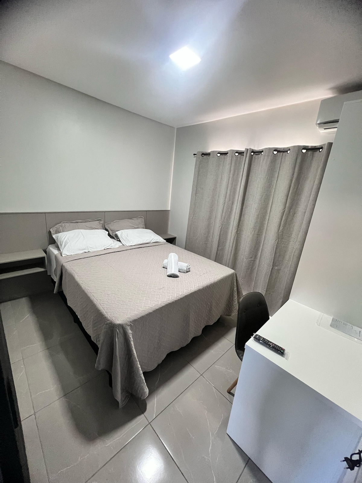 The room features a spacious bed with light bedding, positioned next to a window covered by neutral curtains. A small chair and a desk are present, providing a functional workspace. The bright, clean space is complemented by a smooth tile floor and wall-mounted air conditioning.