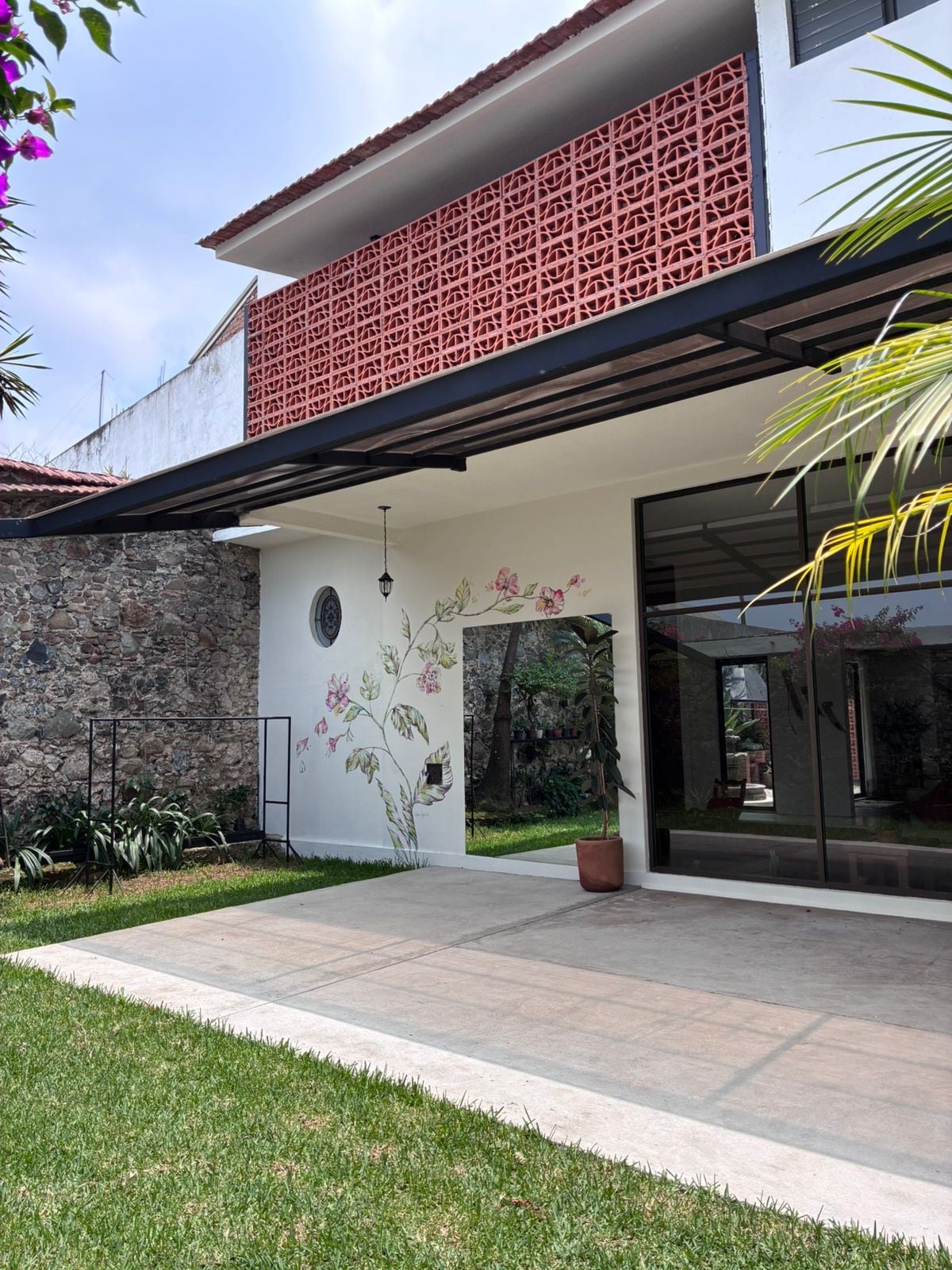 A bright outdoor area is shown, featuring a smooth concrete patio surrounded by lush green grass. Decorative plants are positioned near the edges, while a wall adorned with delicate floral motifs complements the modern architecture of the house. Large glass doors offer a view of the interior space.