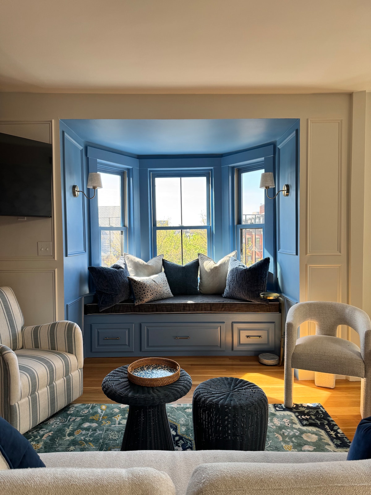 A cozy bay window nook is accented by blue walls and decorative cushions in various textures. Soft lighting from wall sconces enhances the inviting space. Two stylish chairs are positioned nearby, complementing a detailed area rug that adds warmth to the wooden floor.