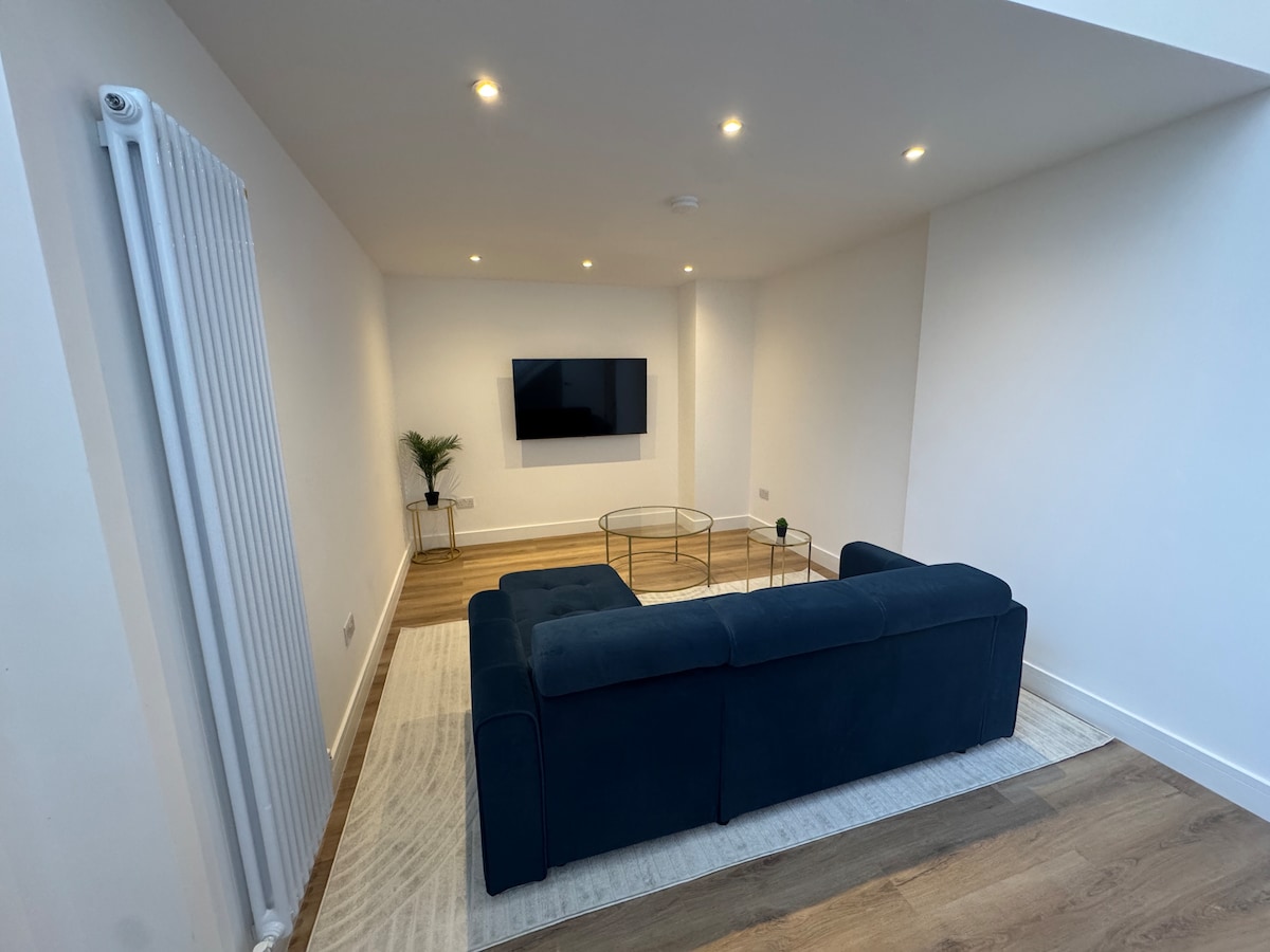 A modern living area features a sleek blue sofa facing a wall-mounted television. A glass coffee table sits on a neutral-toned rug, complemented by a small decorative plant in the corner. Recessed lighting illuminates the space, enhancing its clean design.
