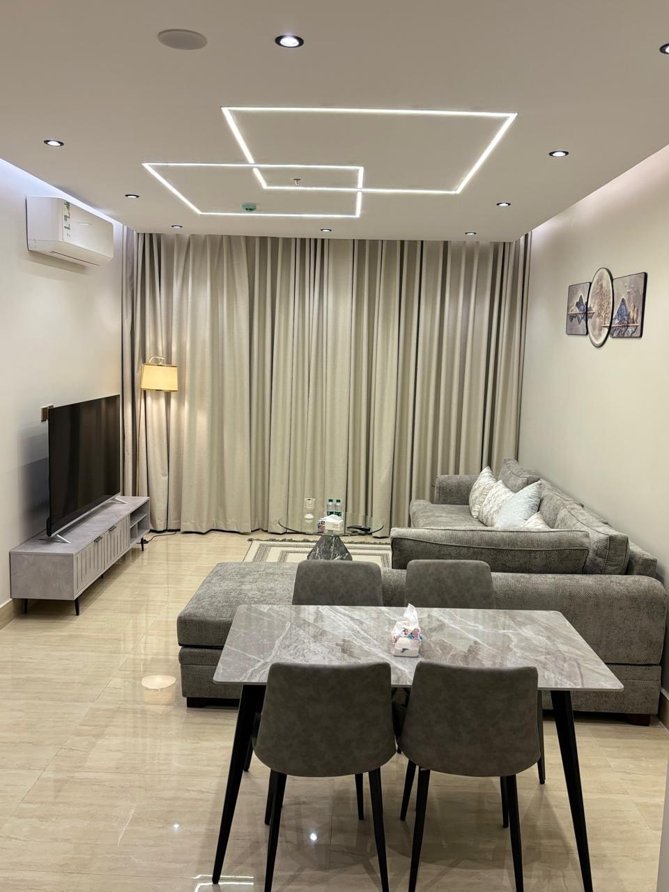 A modern living area features a plush gray sofa paired with matching armchairs, set against softly draped curtains. A sleek entertainment unit supports a flat-screen TV, while a marble dining table with stylish chairs occupies the center of the room, illuminated by recessed lighting.