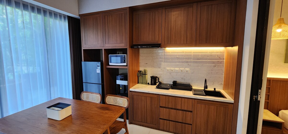 A modern kitchen area features wooden cabinetry and a contrasting white countertop. Essential appliances, including a refrigerator, microwave, and coffee maker, are visible. A dining table with four chairs occupies the space, complemented by large windows allowing natural light to enter.