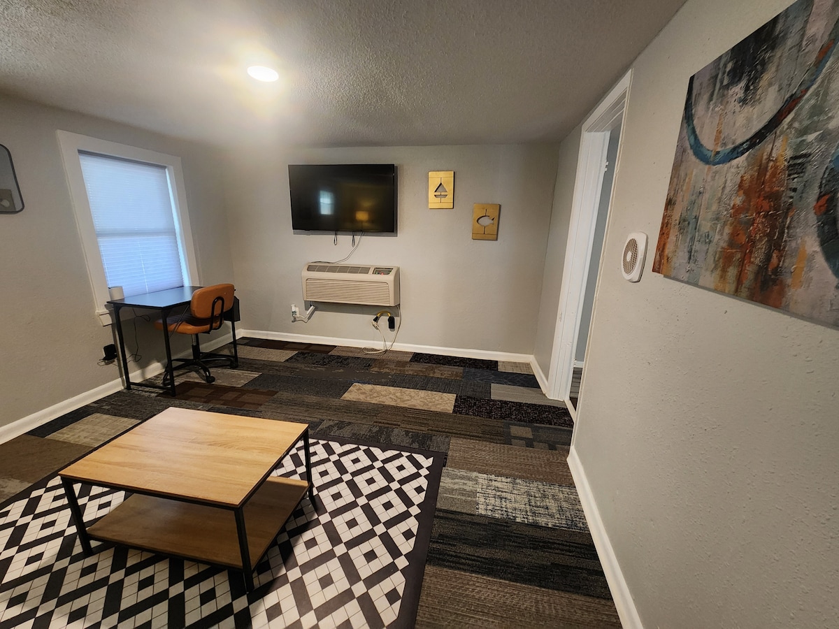 A well-lit living area features a flat-screen television mounted on the wall. A small wooden table is positioned on a patterned rug, accompanied by a chair beneath a window. Abstract art and decorative wall pieces add visual interest to the neutral-toned walls.