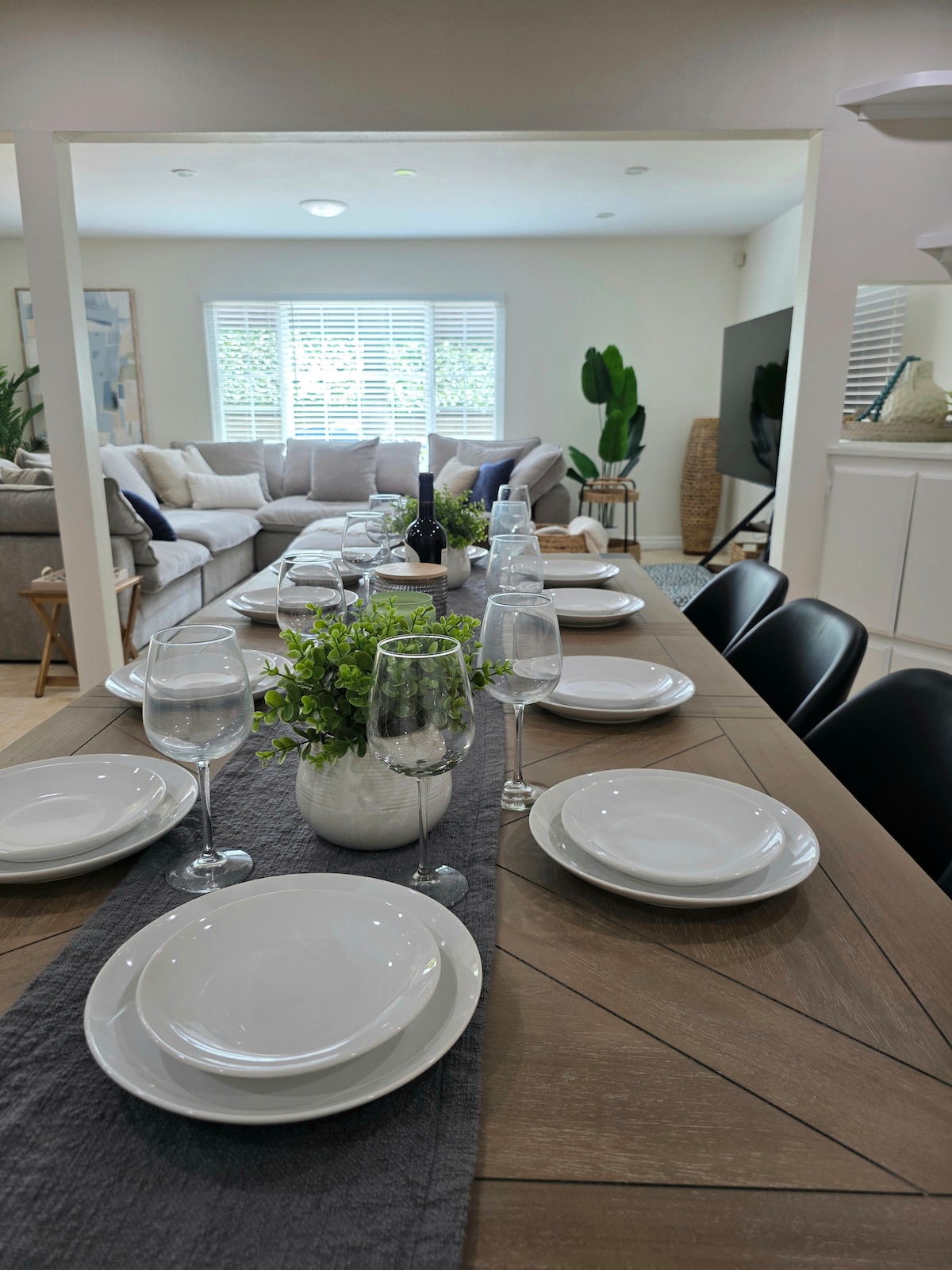 A spacious dining table is set with white plates and glasses, ready to accommodate up to ten guests. A decorative centerpiece featuring a small plant adds a touch of greenery. In the background, a comfortable living area with a large sofa and windows enhances the inviting atmosphere.