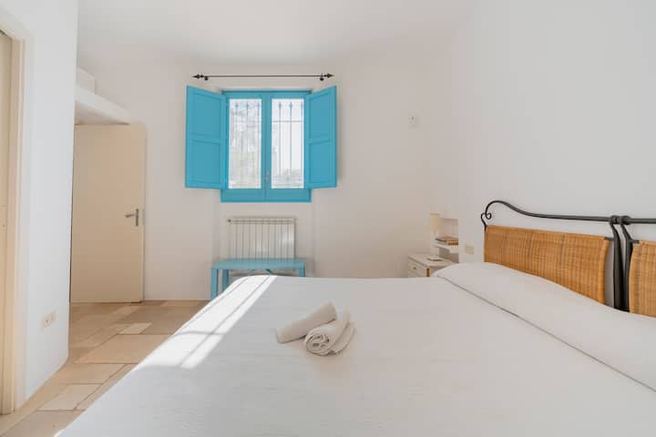 Camera da letto matrimoniale luminosa con pavimento in pietra locale, biancheria da letto confortevole e caratteristiche persiane blu.