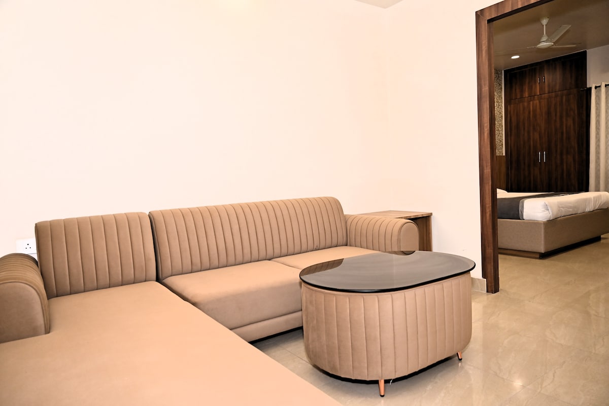 A cozy living area features a ribbed sectional sofa in soft beige tones, complemented by a circular coffee table with a dark surface. A light marble floor enhances the space, and a faint glimpse of a bed is visible in the background, adding to the comfort of the area.