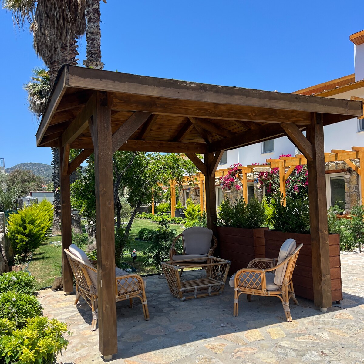 A wooden gazebo provides a shaded seating area surrounded by greenery and flowering plants. Four wicker-style chairs are positioned around a small table, creating a welcoming space for relaxation. Sunlight filters through the open structure, illuminating the nearby garden.