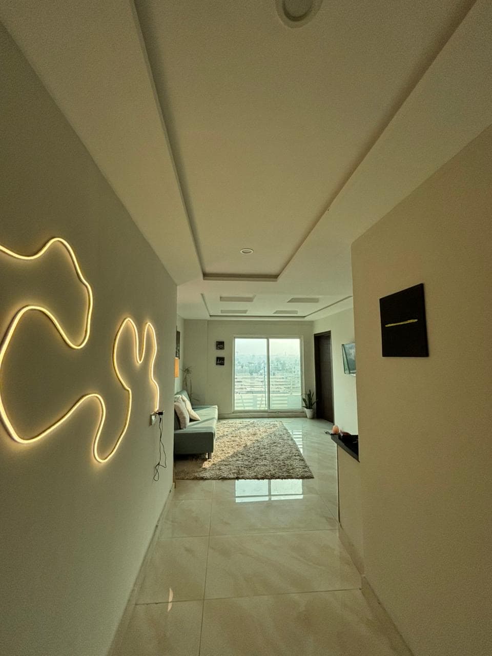 A hallway leads into a bright living space, featuring smooth, light-colored walls and glossy tile flooring. A framed artwork hangs near the door, while a modern light fixture contrasts with the decor. Large glass doors provide views of the outside, enhancing natural light.