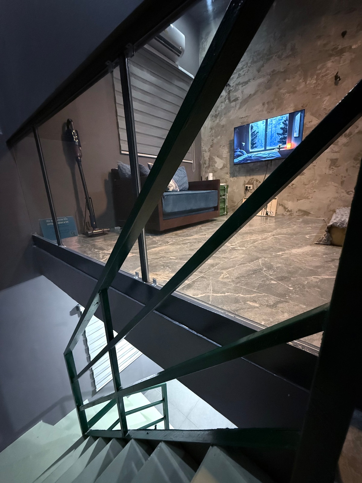 A modern staircase with green railings leads down to a living area featuring a dark sofa and a large television against a textured wall. Soft lighting and a tall lamp create an inviting ambiance in the space.