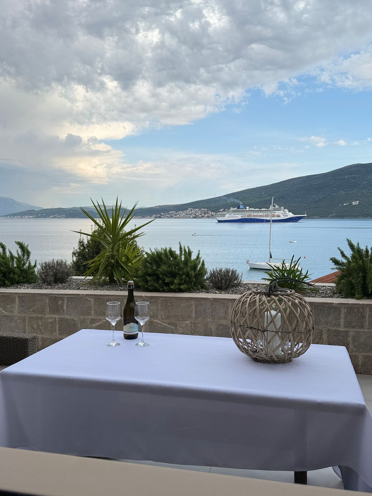 A table draped in a white cloth is set for two, featuring glasses and a bottle. Lush greenery is visible in the foreground, while a scenic view of the water and mountains stretches out in the background, with a boat passing near the shore.