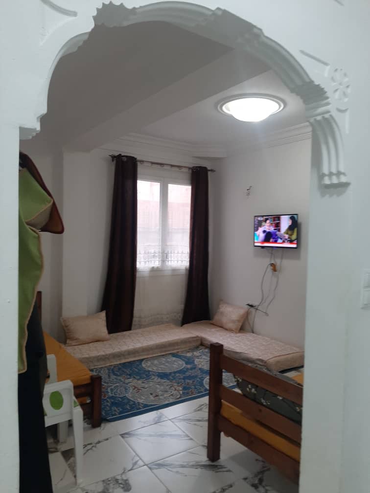 A cozy seating area is visible, featuring floor cushions arranged on a traditional rug. A flat-screen television is mounted on the wall, while natural light enters through a window adorned with dark curtains. The room is framed by an arched entrance, enhancing the space.