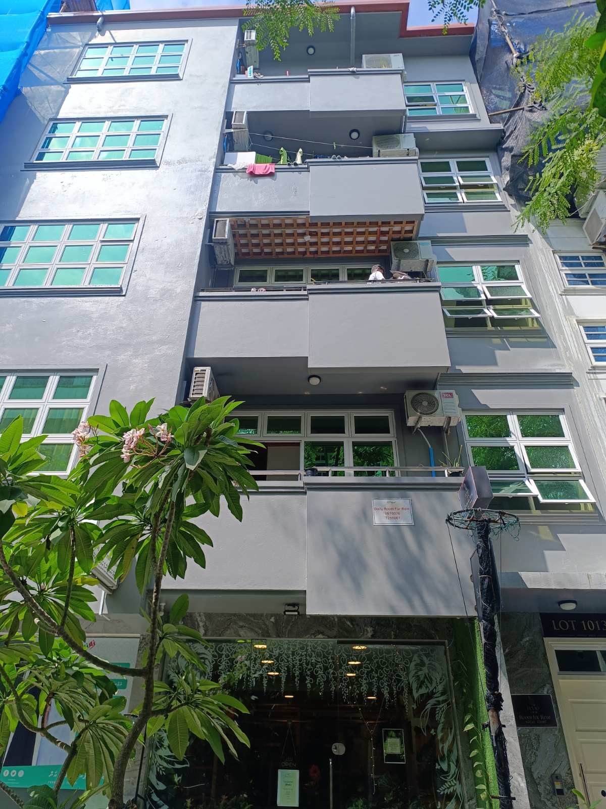 The exterior of the building is captured from below, showcasing a modern, multi-story structure with large green-framed windows. A balcony with wooden slats is visible on the second floor, while plants add a touch of greenery to the surroundings.