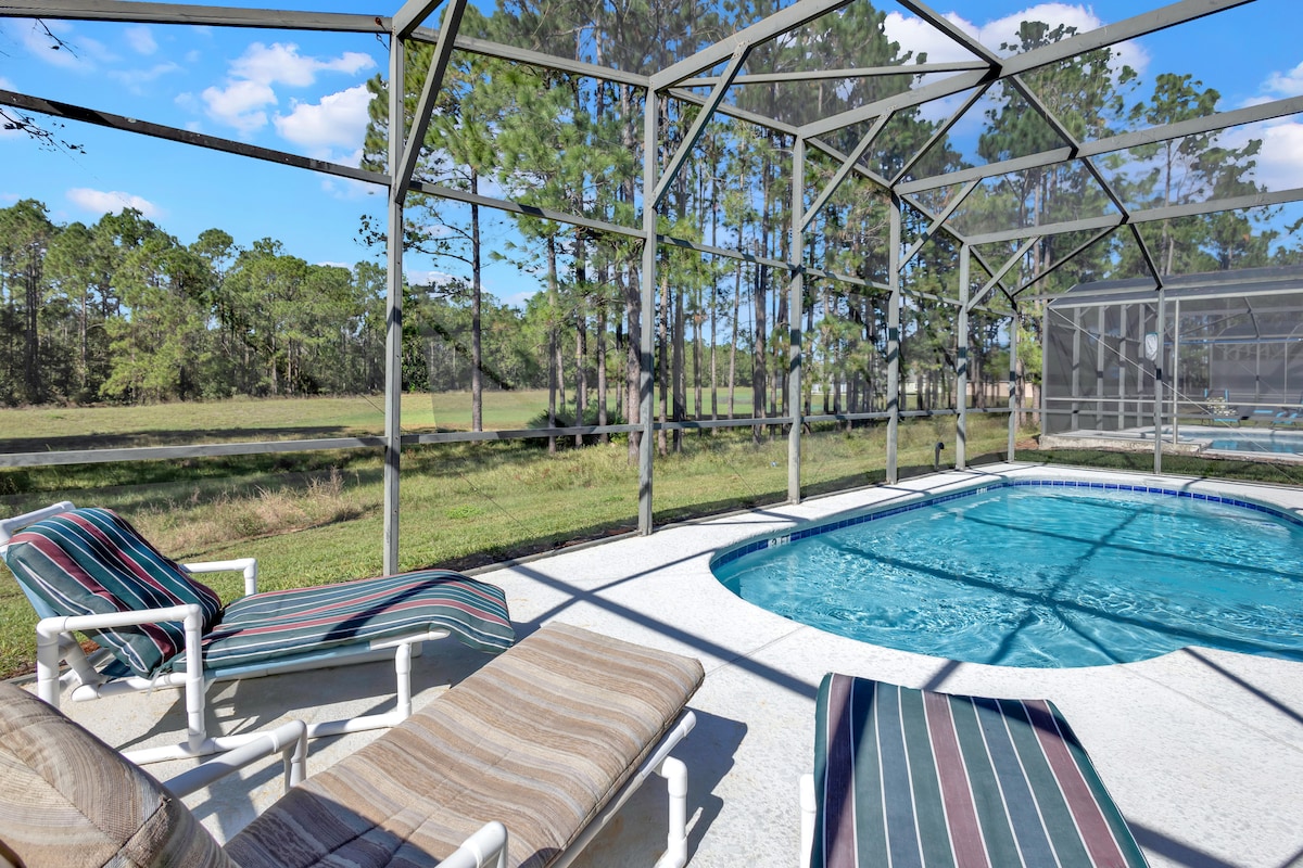 An enclosed pool area features a clear blue pool surrounded by a spacious deck. Two lounge chairs are positioned beside the pool, and a backdrop of tall trees provides privacy. Sunlight filters through the screened enclosure, creating a bright and fresh ambiance.