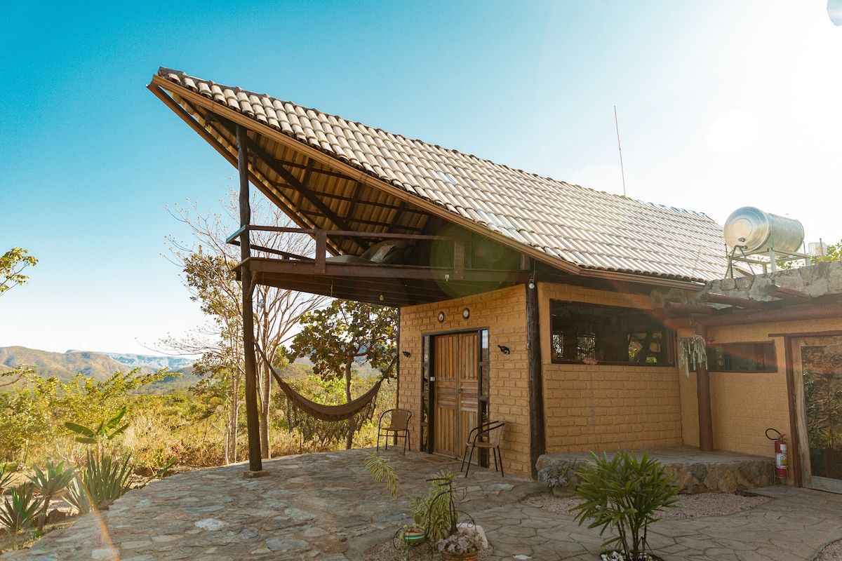 A charming eco-lodge sits with an angled roof and a spacious outdoor area. Comfortable chairs are positioned on the stone patio, inviting relaxation. Lush vegetation and distant mountains provide a picturesque backdrop under a clear blue sky.