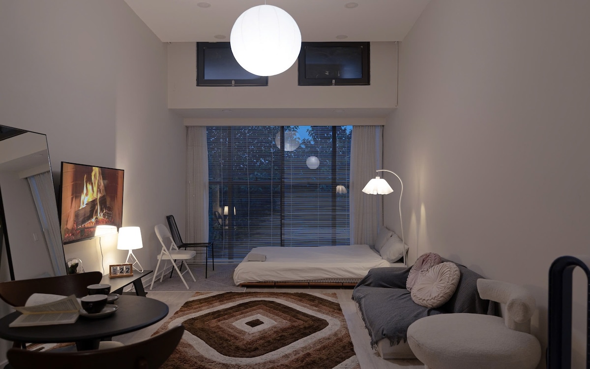 A well-lit living space is complemented by large windows that allow natural light to flow in. A spacious bed is neatly positioned on one side, and a cozy seating area featuring a soft rug encourages relaxation. A Smart TV is mounted above a dark table set for dining.