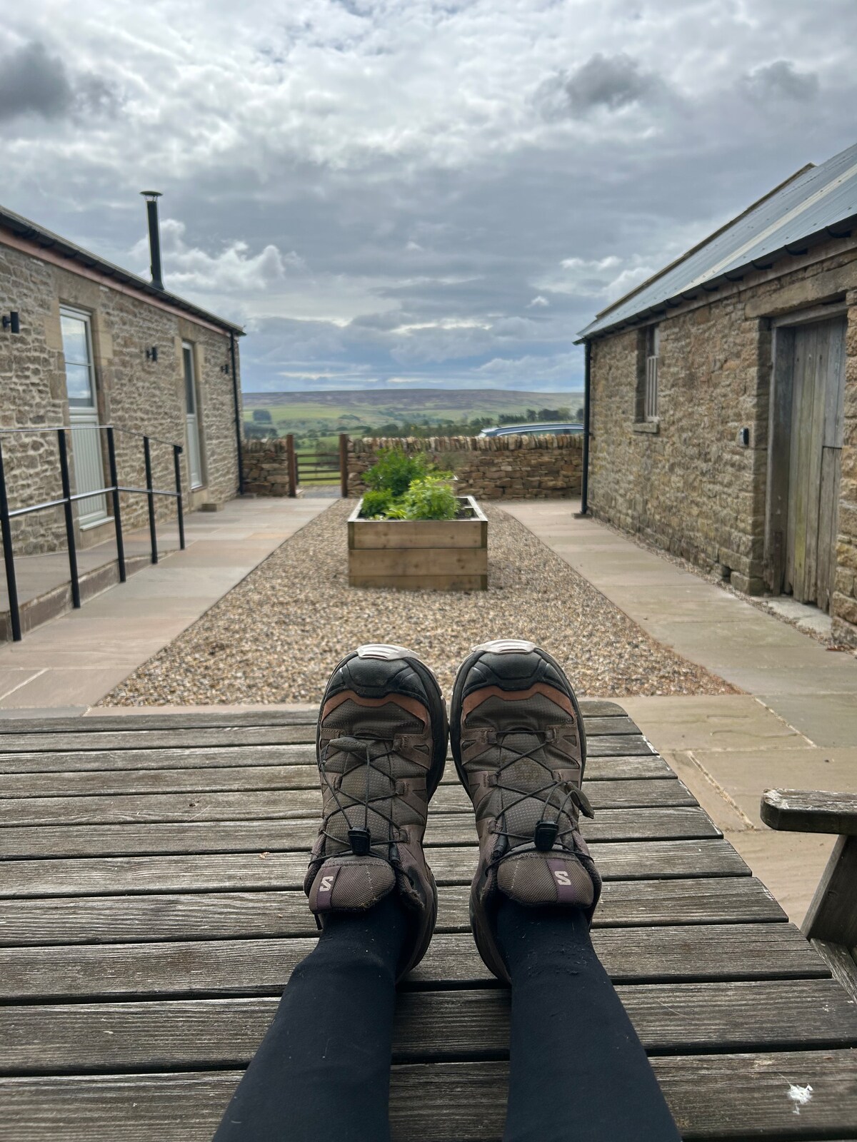 A pair of hiking shoes rests on a wooden table, with legs extending toward a gravel pathway. Surrounding the path are stone buildings and a landscaped planter filled with greenery. Above, a partly cloudy sky stretches over scenic countryside views.