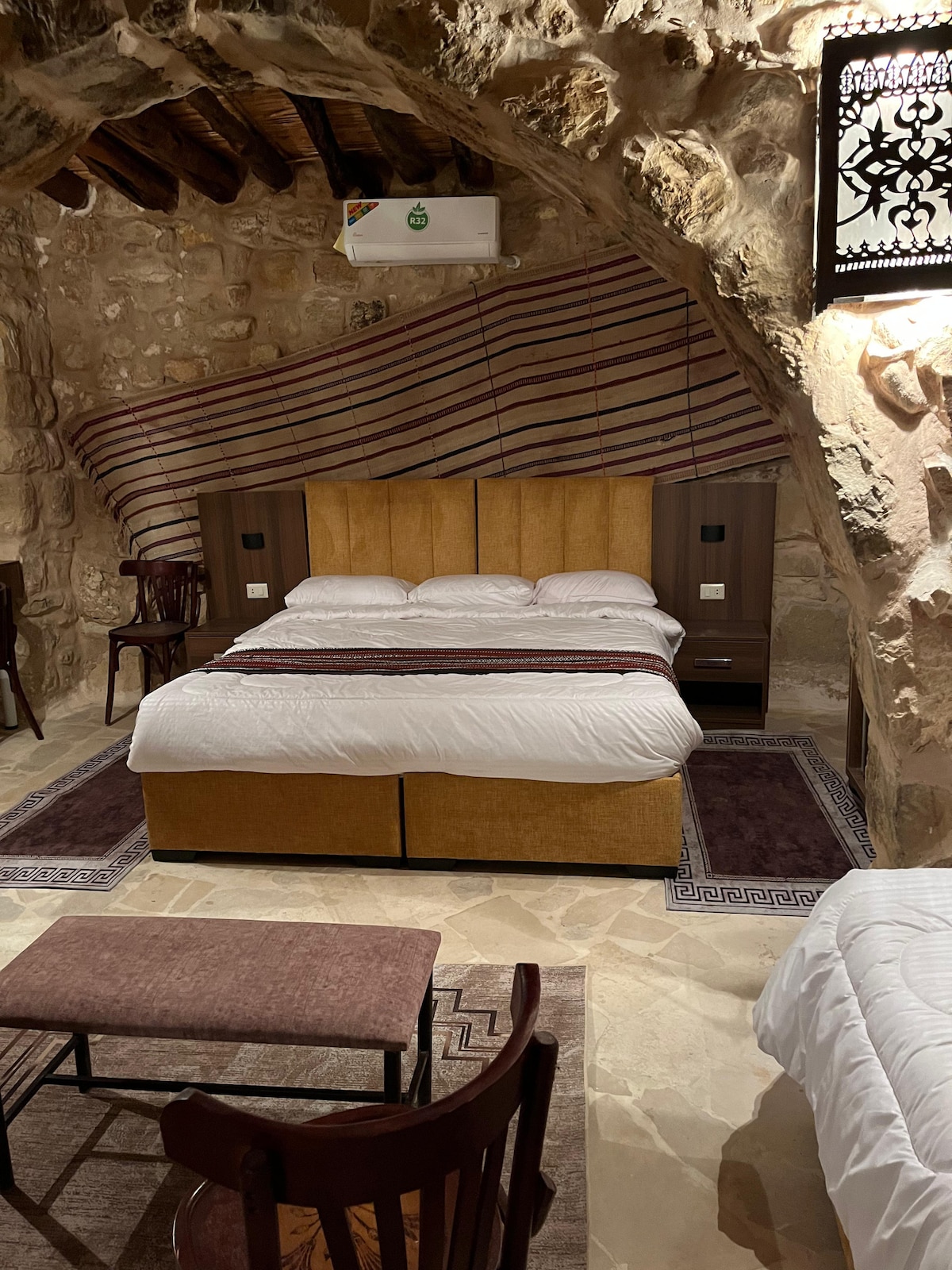 A spacious room with a large bed dressed in white linens is framed by rustic stone walls. Traditional textiles hang above the bed, adding cultural detail. A wooden table and chairs are arranged nearby, offering a cozy seating area, while soft lighting creates a calming atmosphere.