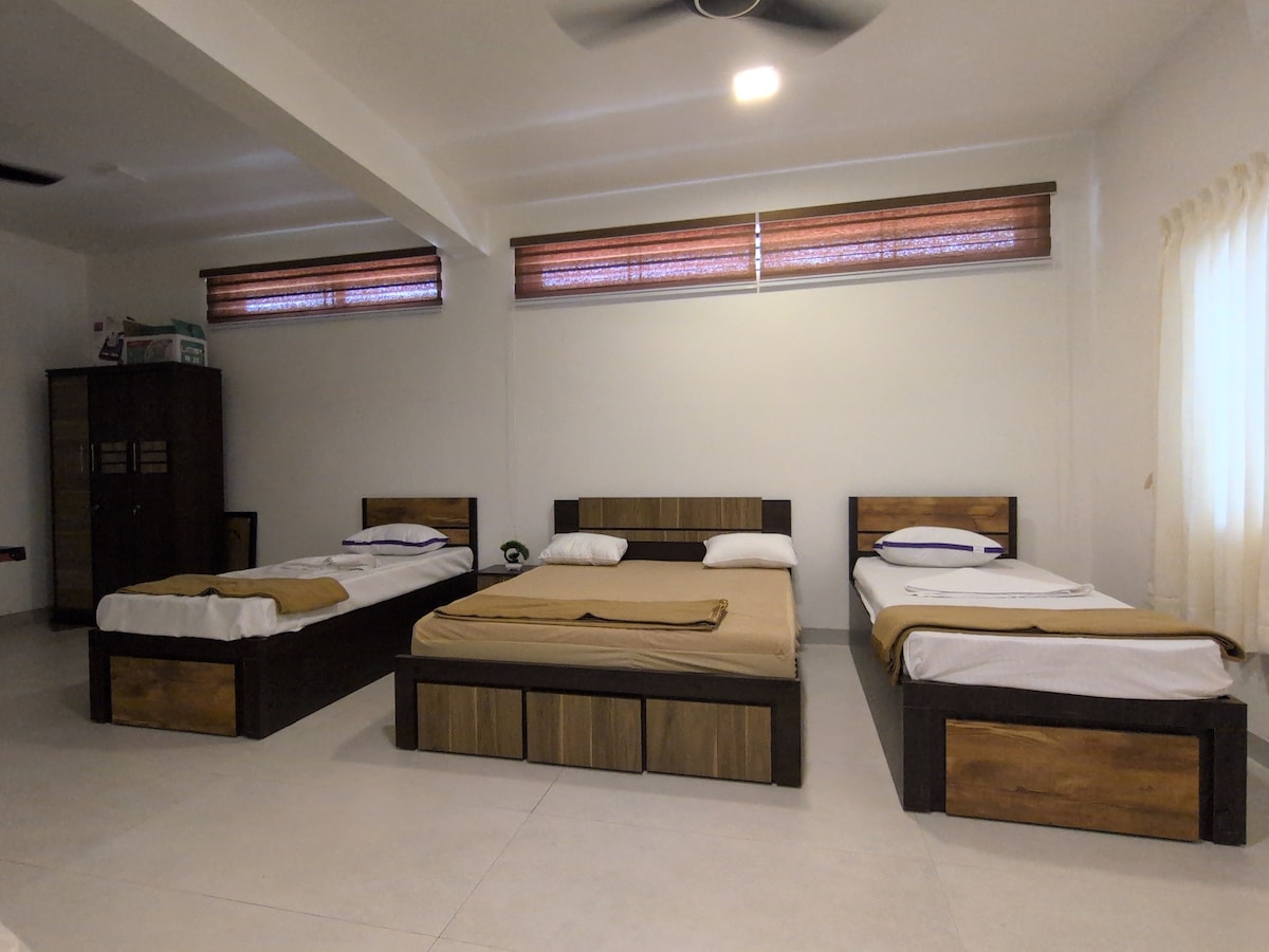 The sleeping area contains a spacious queen bed and two single beds, arranged to accommodate multiple guests. Natural light enters through two windows with wooden blinds, and a dark storage unit is positioned against the wall, providing additional space for belongings.