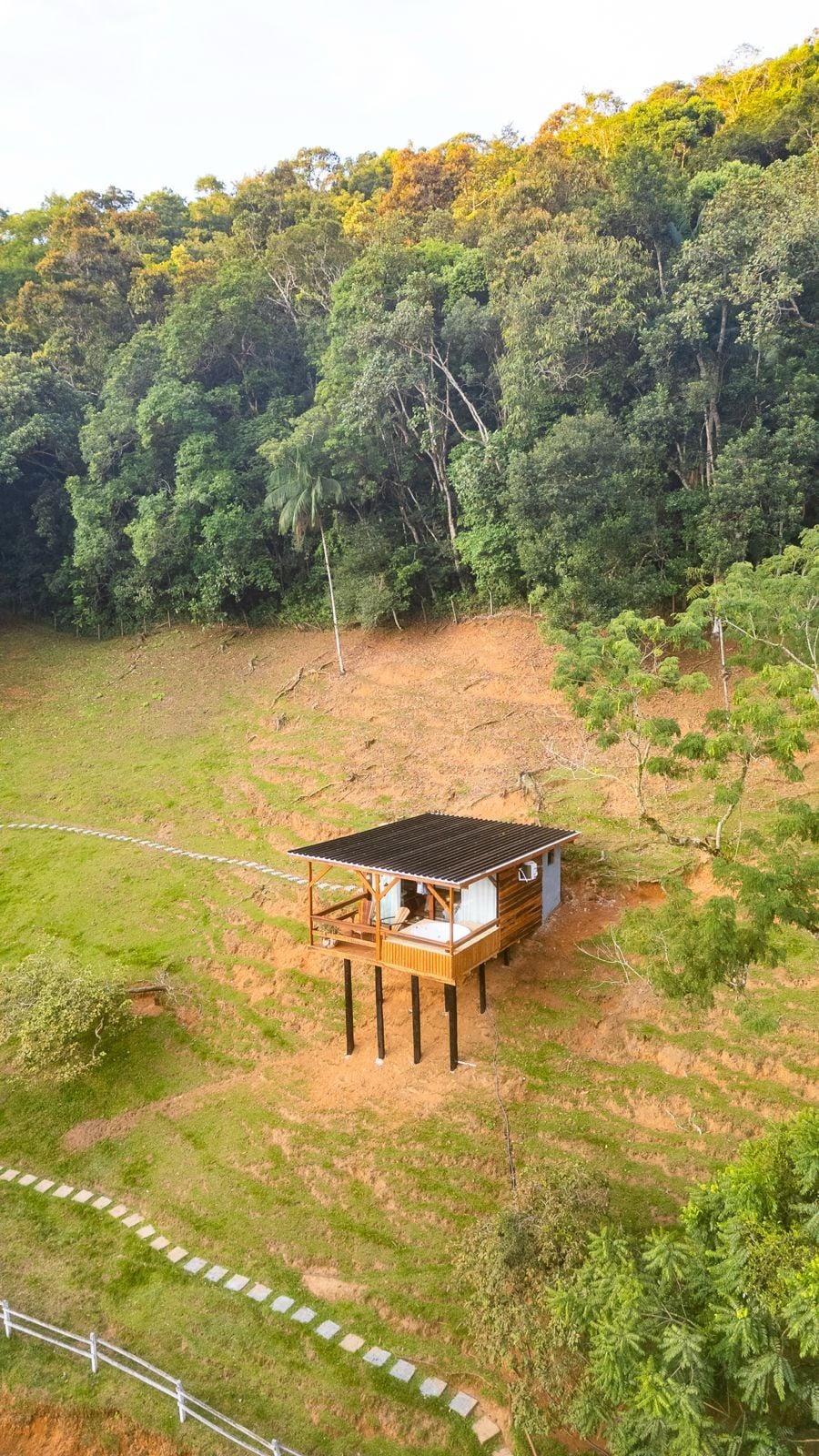 A charming cabana, elevated on stilts, is nestled within lush greenery. Surrounded by trees, a perspective reveals the sloping terrain below. The structure features a covered porch, providing an outdoor space to enjoy the natural surroundings.