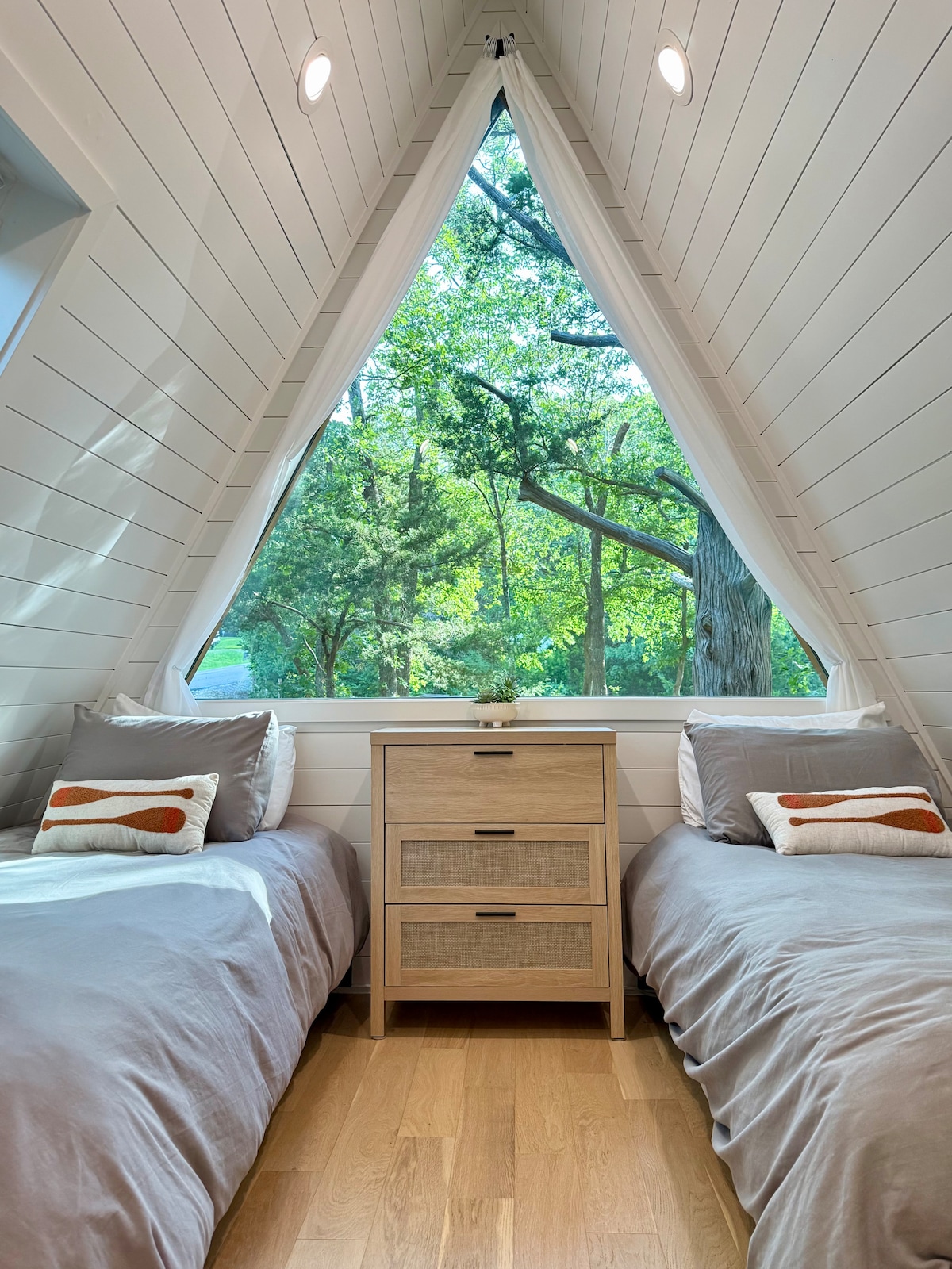 A cozy bedroom features two twin beds, each adorned with soft grey bedding and decorative pillows. A simple dresser stands between the beds, while large windows framed in wood offer a view of the surrounding greenery, creating a serene atmosphere.