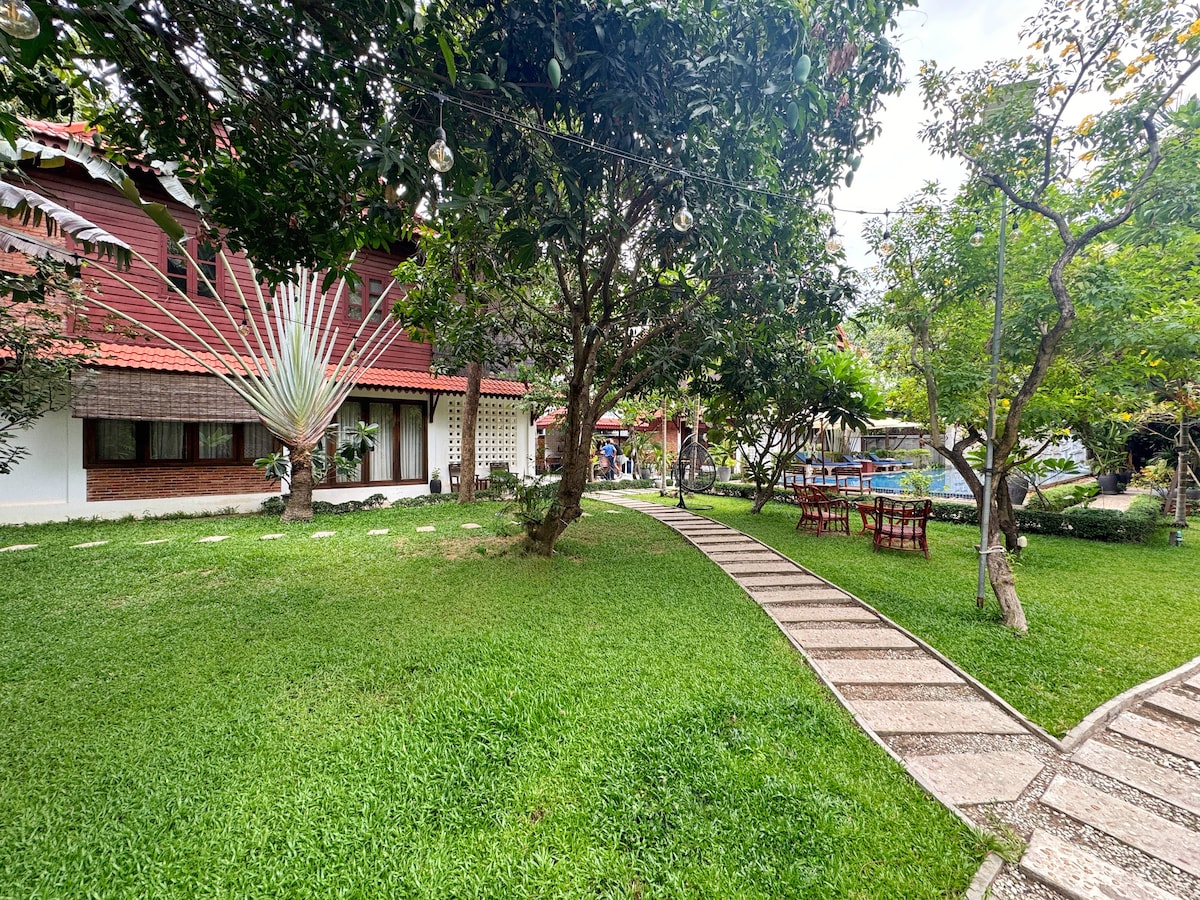 A lush green garden area is featured, with smooth stone pathways winding through manicured grass. Surrounding trees provide shade, and colorful plants add natural beauty. A seating area with a table and chairs can be seen, alongside various outdoor decorations, creating a relaxed outdoor atmosphere.