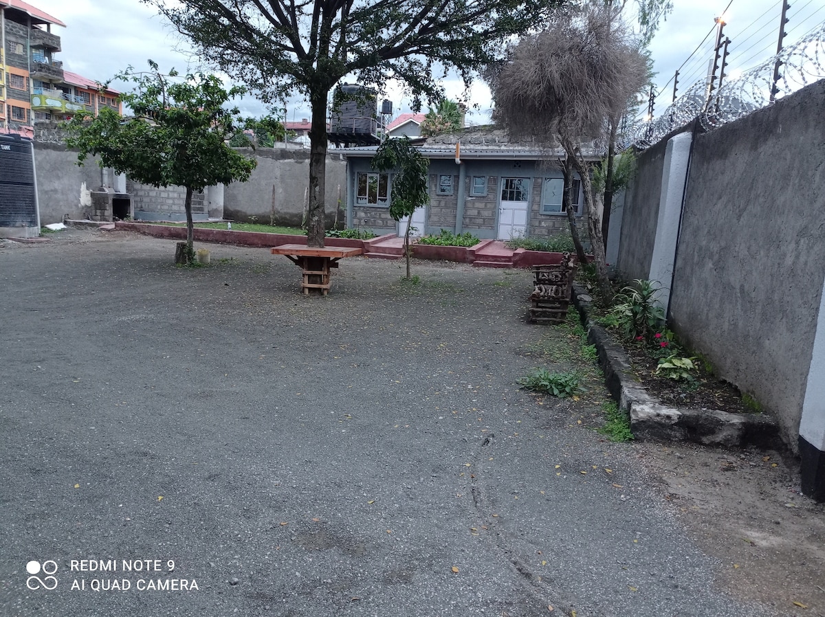 An outdoor area is shown with gravel flooring and surrounding walls. A wooden table with benches is situated in the center, while a small garden with plants features on the right side. Trees provide shade, and a building is visible in the background.