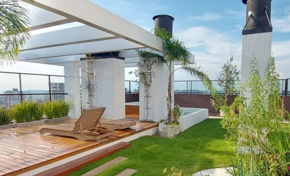 A rooftop area is featured with wooden decking, lush green grass, and designated spaces for relaxation. A sun lounger is positioned beside a minimalist tub, and vertical gardens adorn the walls, complementing the urban backdrop and open sky.