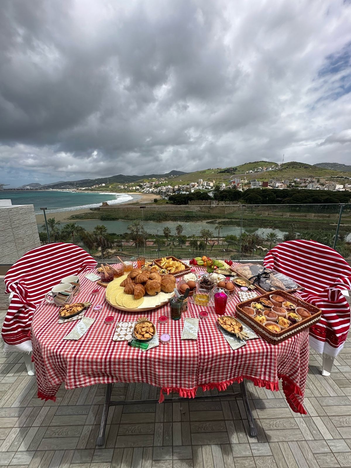 A dining table is set on a terrace, draped with a red and white checkered tablecloth. An array of dishes is displayed, including baked goods and colorful beverages. The background features a scenic view of the sea and hills, under a cloudy sky.