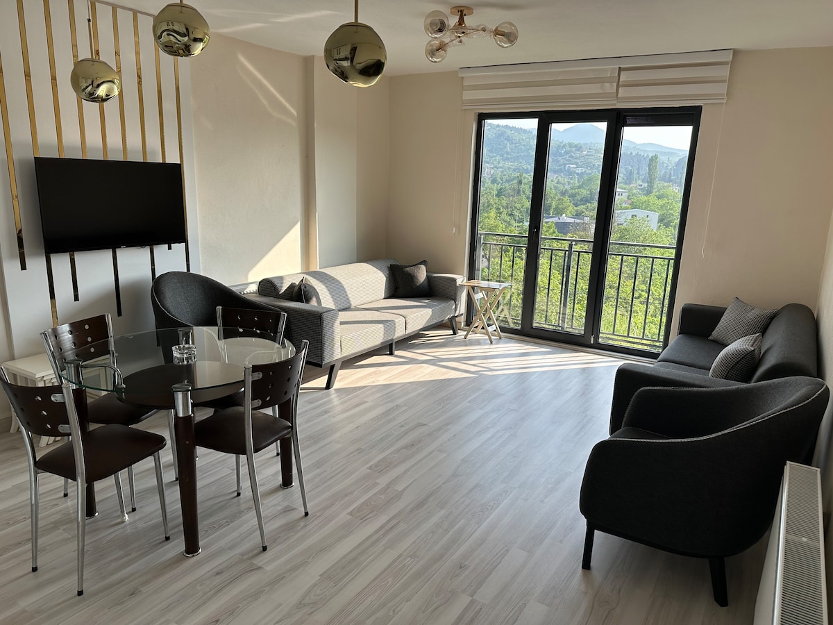A bright living space features large glass doors that open to views of greenery. A glass dining table is surrounded by metallic chairs, while two dark sofas and an armchair provide seating. A wall-mounted television is positioned above a modern light fixture.