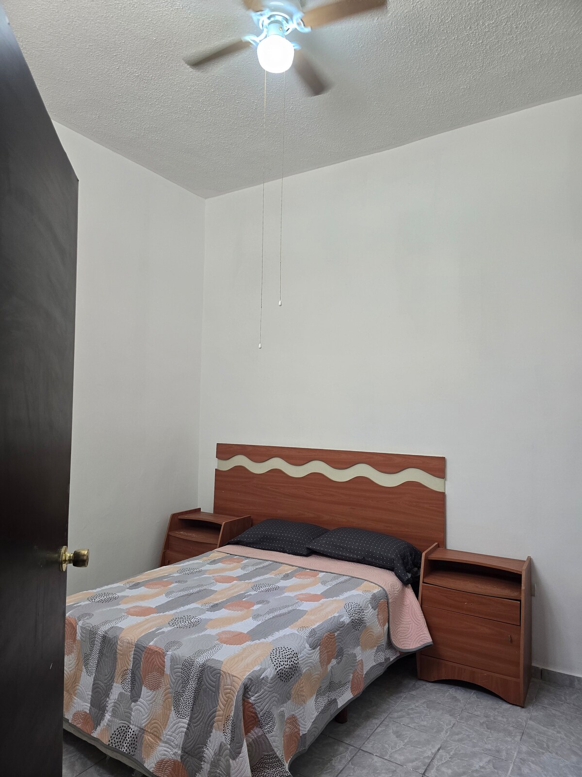 A cozy sleeping area features a single bed with a patterned bedspread and a wooden headboard. Two bedside tables are positioned on either side of the bed. A ceiling fan adds airflow, while natural light enters through a nearby door.