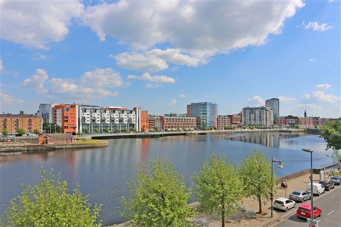 A scenic view of a waterfront area is presented, highlighting a calm river reflecting the blue sky and clouds. Various modern buildings line the opposite shore, surrounded by greenery. A tree-lined promenade is visible in the foreground, enhancing the inviting atmosphere of the area.