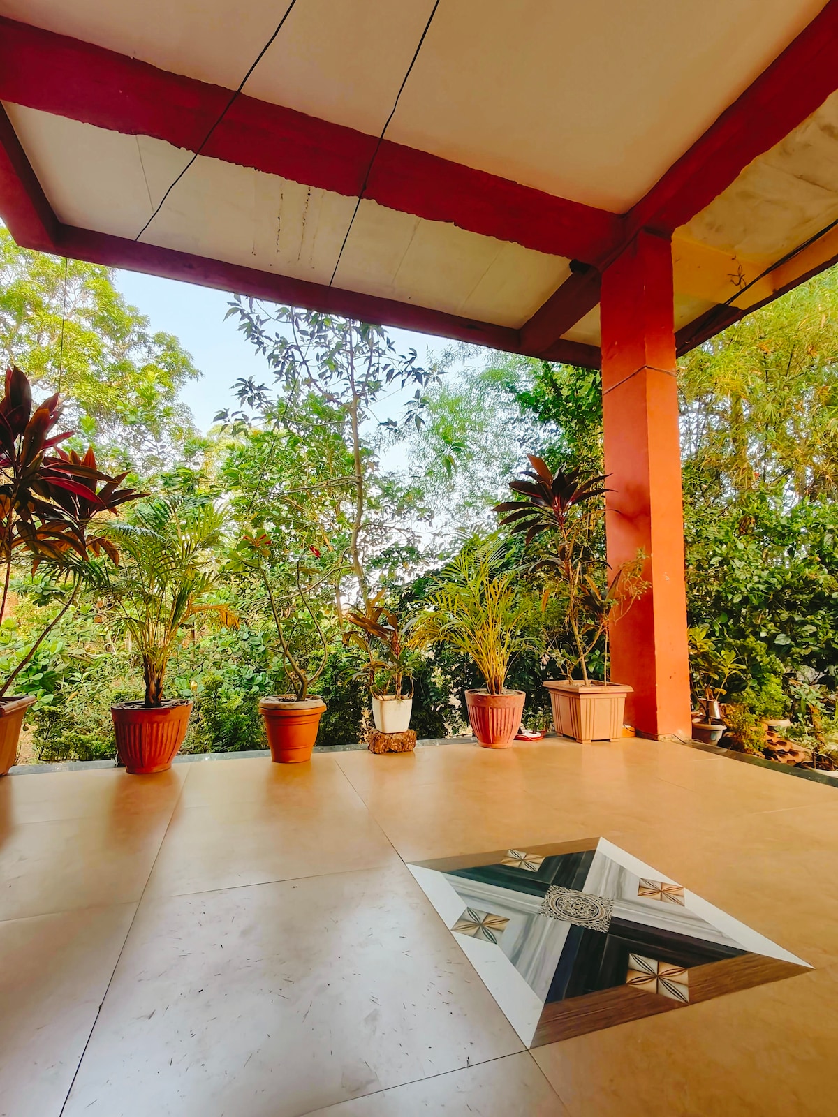 An open patio space is highlighted by several potted plants arranged along the edges. The flooring features an intricate design, and overhead, beams in a red hue support the ceiling while natural light filters through the surrounding greenery.
