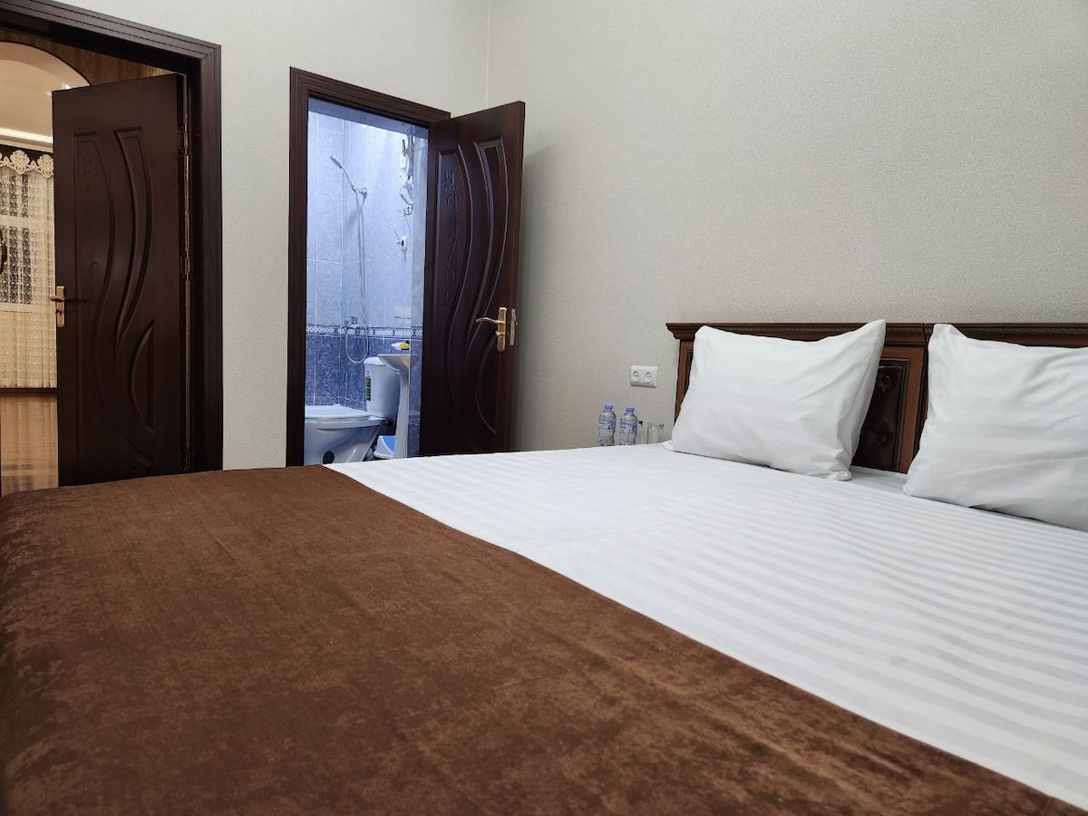 A cozy bedroom features a spacious bed dressed in a neutral-colored blanket. Two light-colored pillows are neatly arranged at the head of the bed. An open door leads to a bathroom area, subtly hinting at the room's layout.