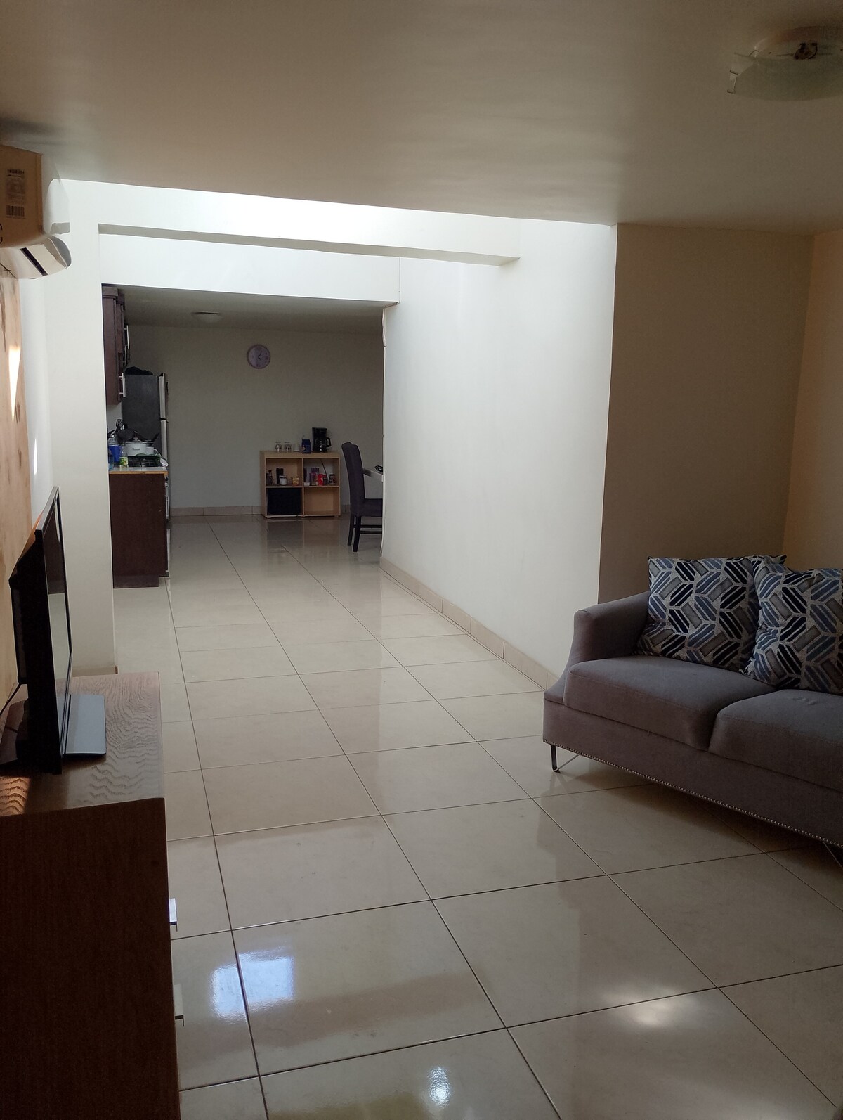A bright and open interior space features glossy tiled flooring illuminated by natural light. A small sofa with patterned cushions is positioned near a television. A dining area with chairs and a kitchen can be seen in the background, creating a connected living environment.