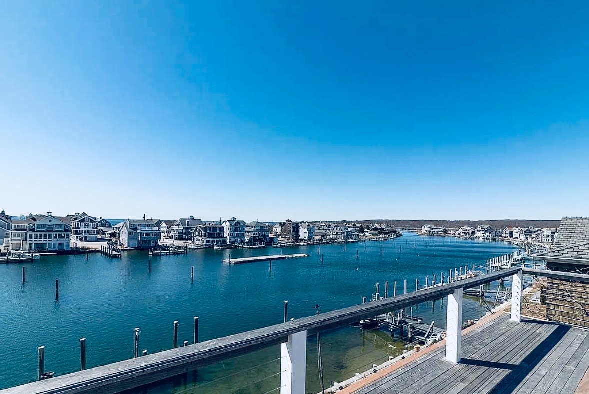A sweeping view of a serene waterway is captured from a private sun-bedding deck. The calm water is lined with charming homes, docks, and boats, set against a clear blue sky. The scene conveys a tranquil coastal atmosphere.